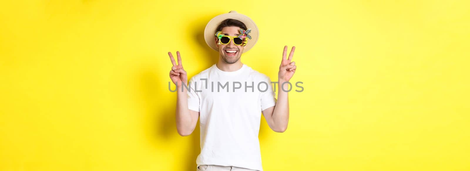 Concept of tourism and lifestyle. Happy man enjoying trip, wearing summer hat and sunglasses, posing with peace signs for photo, yellow background.