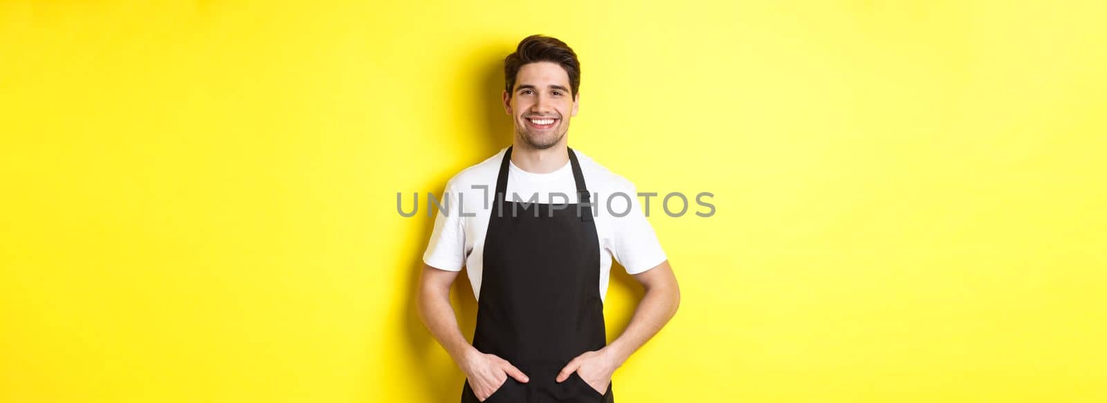 Happy barista in black apron looking at camera. Coffee shop owner wearing cafe uniform and smiling, standing over yellow background.