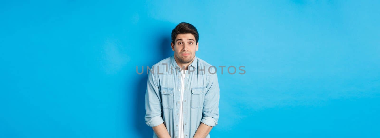 Embarrassed guy want to pee, waiting in line for toilet, standing against blue background.