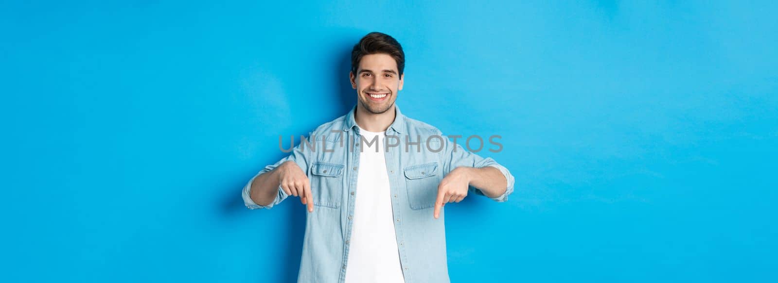 Handsome smiling adult man introduce product, pointing fingers down at promotion, standing against blue background.