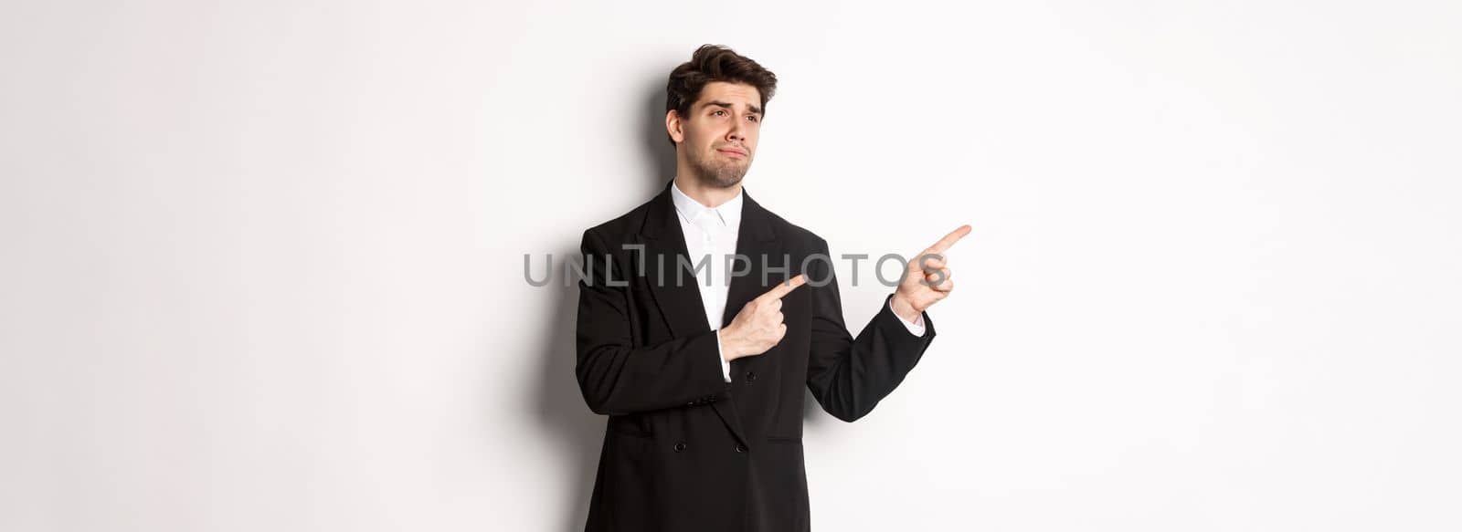 Image of upset and disappointed handsome guy in formal suit, pointing and looking left with sad face, standing over white background by Benzoix