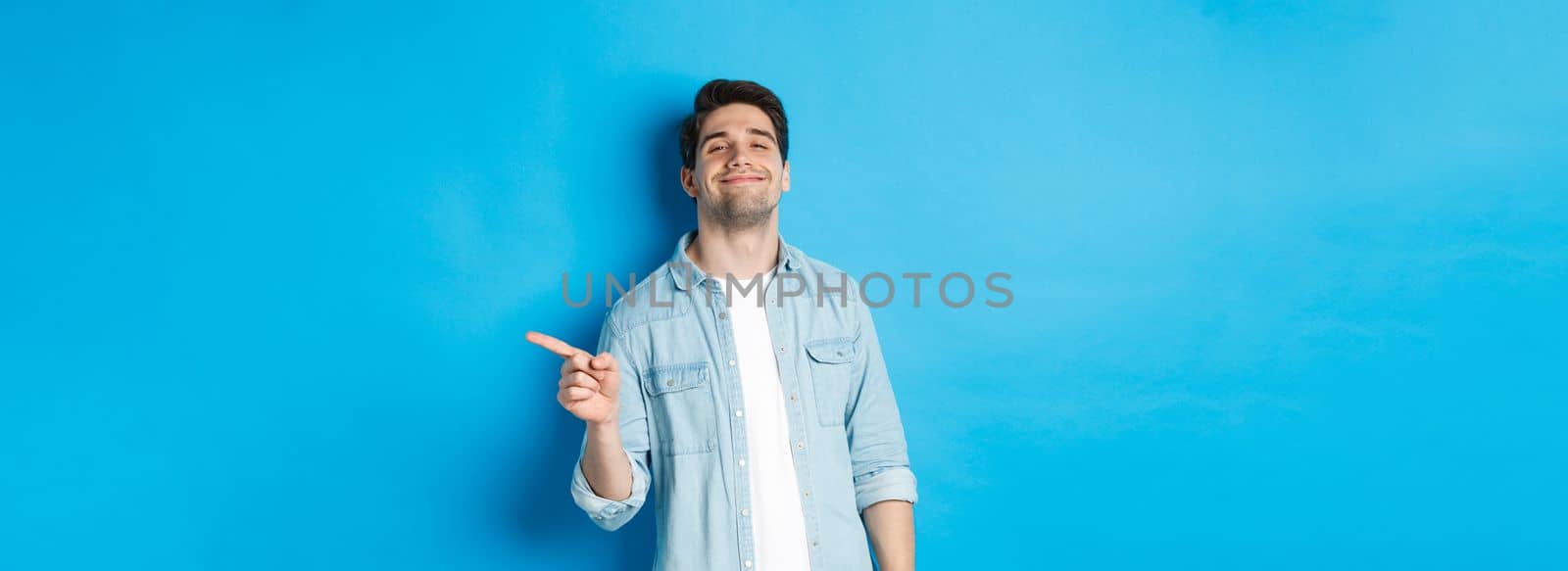 Confident handsome guy smiling sassy, pointing finger right at copy space, showing advertisement, standing over blue background.