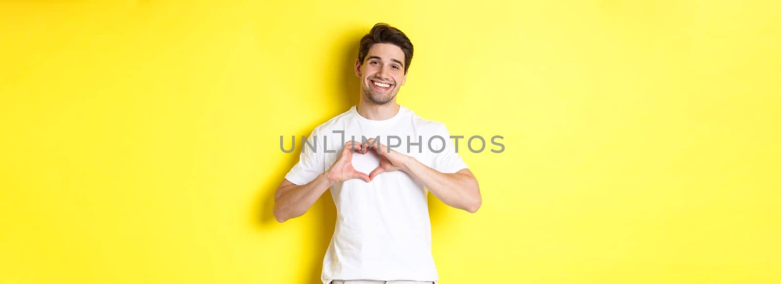 Happy romantic man showing heart sign, smiling and express love, standing over yellow background by Benzoix