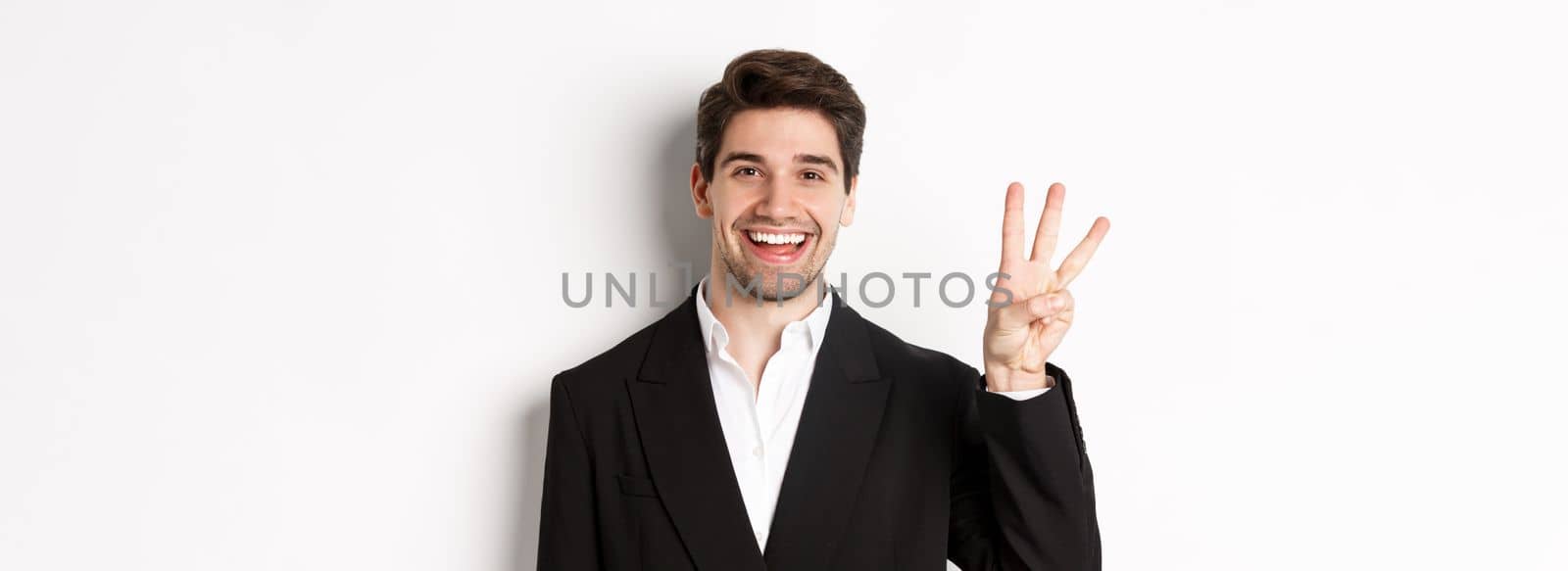 Close-up of handsome businessman in black suit, smiling amazed, showing number three, standing over white background by Benzoix