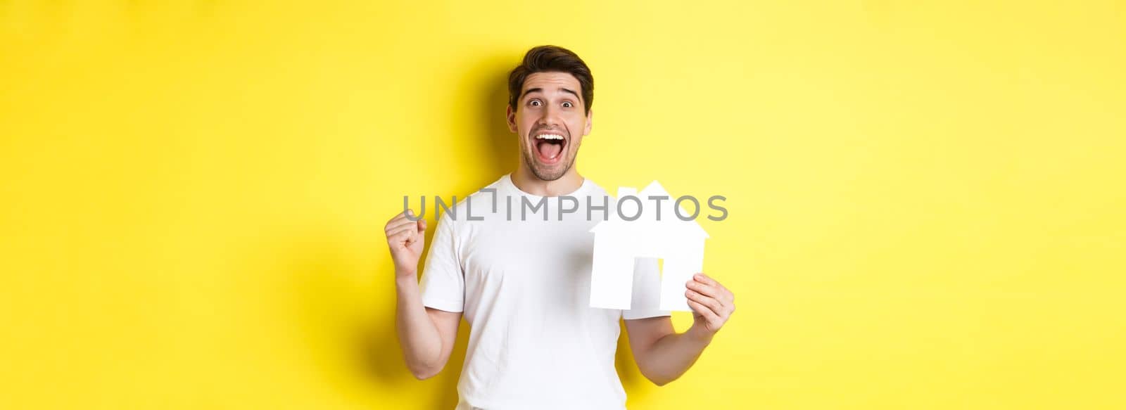 Real estate concept. Excited man holding paper house model and celebrating, standing happy over yellow background by Benzoix