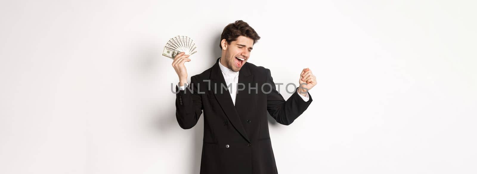 Portrait of handsome and successful businessman in suit, dancing with money, standing against white background by Benzoix