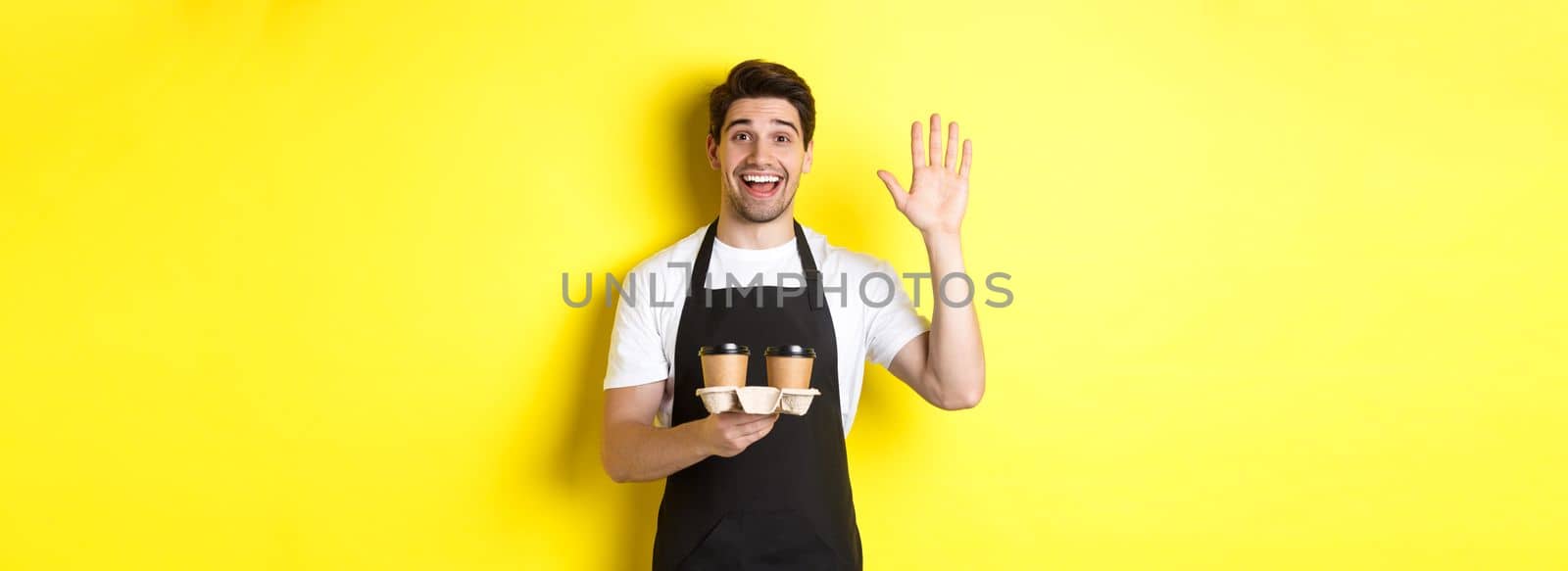Friendly waiter in cafe waving hand at customer, holding takeaway coffee oder, standing against yellow background in black apron by Benzoix