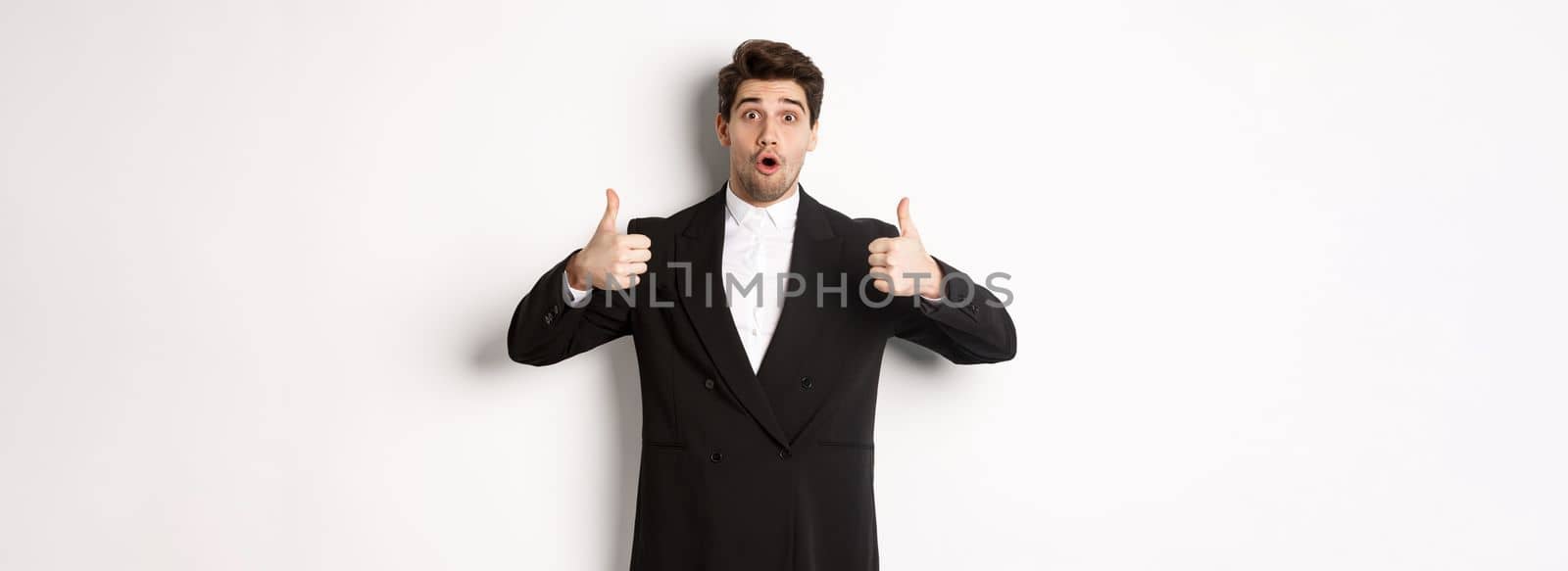 Concept of new year party, celebration and lifestyle. Portrait of excited handsome businessman in formal suit, showing thumbs-up in approval, recommending shop, standing over white background by Benzoix