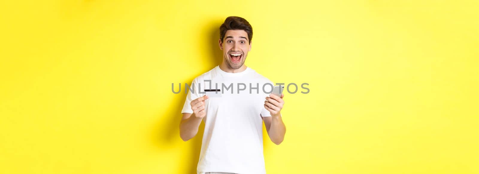 Happy male buyer holding smartphone and credit card, concept of online shopping in internet, standing over yellow background.