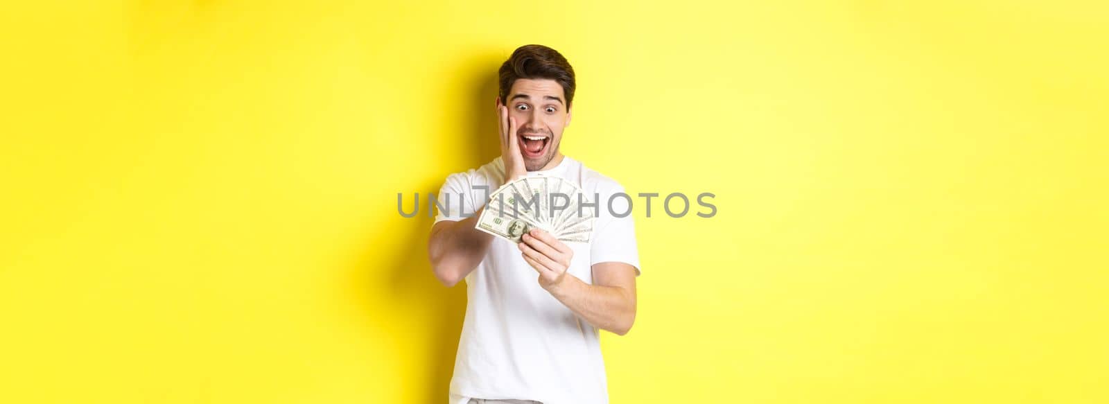 Man looking amazed at money, winning cash prize, standing against yellow background.