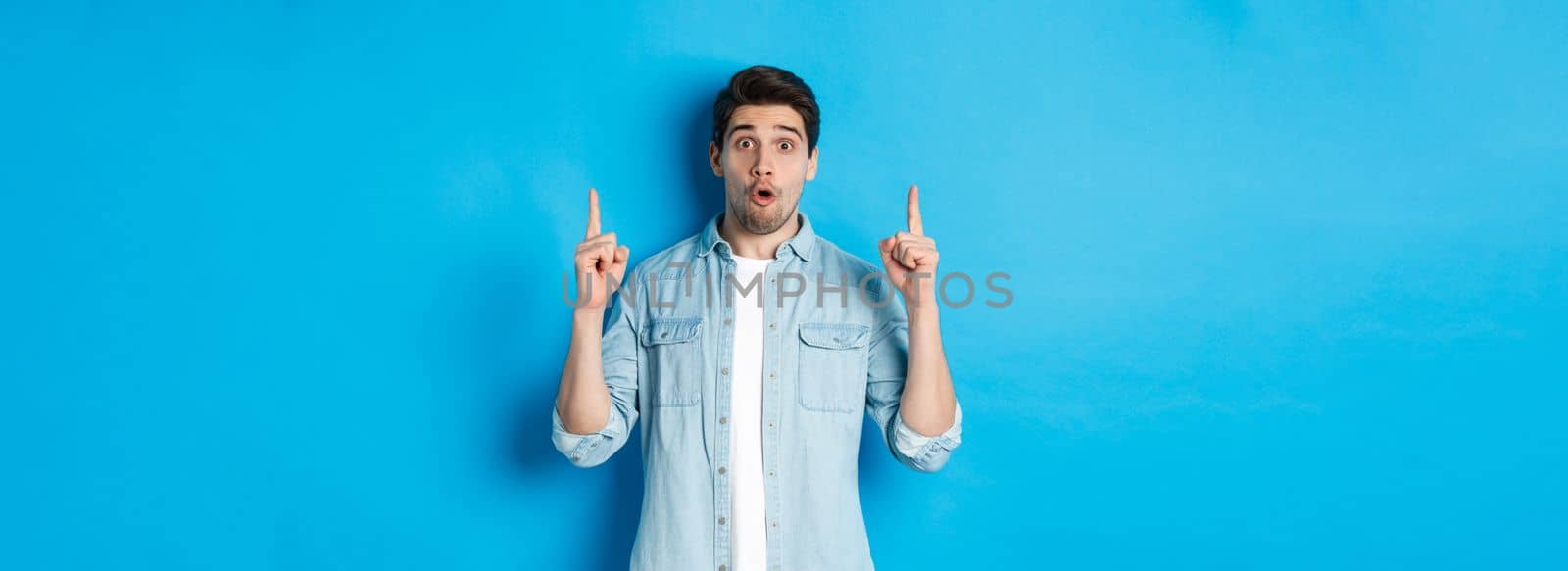 Portrait of amazed handsome man pointing fingers up, showing logo on blue background.