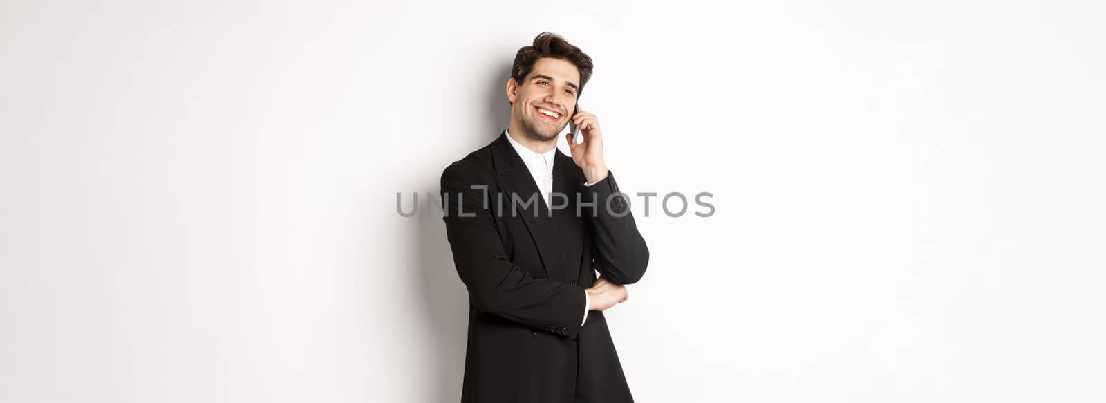 Image of handsome and successful businessman talking on phone, smiling pleased, standing in suit against white background.