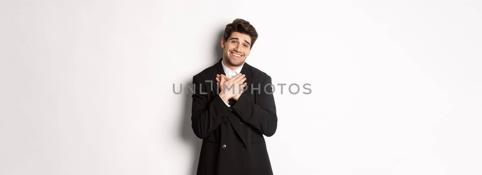 Portrait of happy and pleased handsome man in party suit, holding hands on heart and sighing flattered, saying thank you, standing grateful over white background.