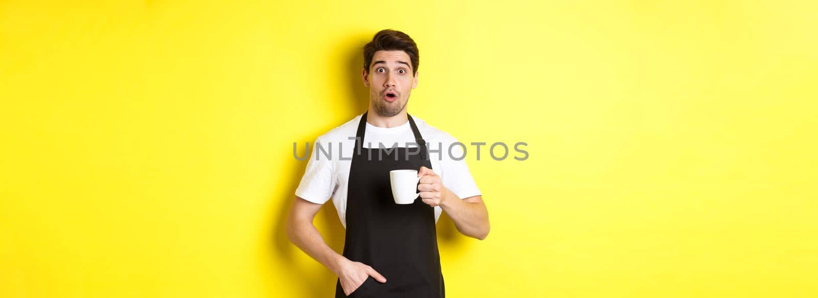 Barista holding coffee mug and looking surprised, standing in black apron cafe uniform against yellow background by Benzoix