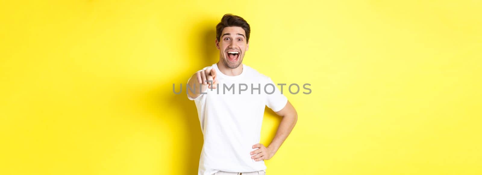 Happy guy pointing finger at camera and laughing, check out something, standing over yellow background by Benzoix