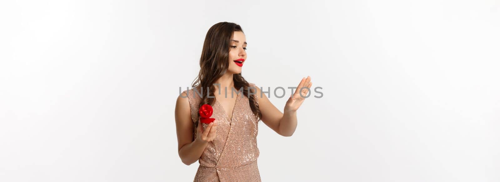 Beautiful woman in luxury dress, trying on engagement ring, saying yes to marriage proposal, looking dreamy at hand, standing over white background.