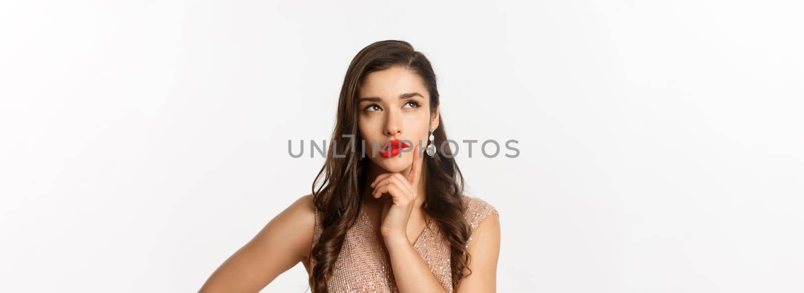 Concept of New Year celebration and winter holidays. Close-up of elegant woman with red lips and dress, looking at upper left corner and thinking, standing over white background by Benzoix