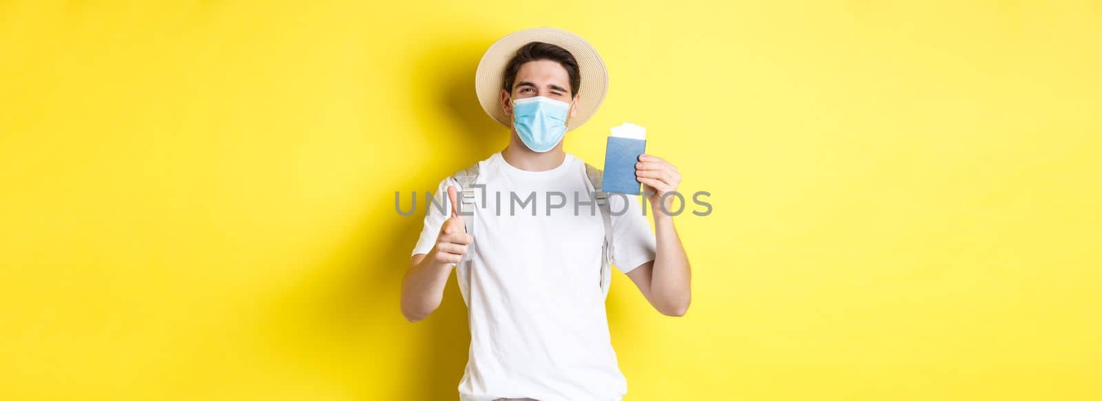 Concept of covid-19, tourism and pandemic. Happy guy tourist in medical mask showing passport with tickets for vacation, winking and pointing at camera, yellow background.