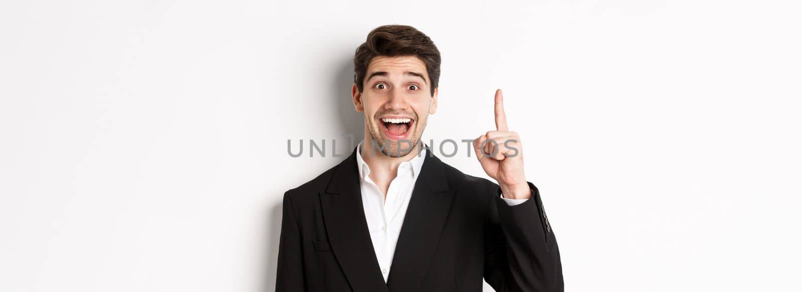 Close-up of handsome businessman in black suit, smiling amazed, showing number one, standing over white background.