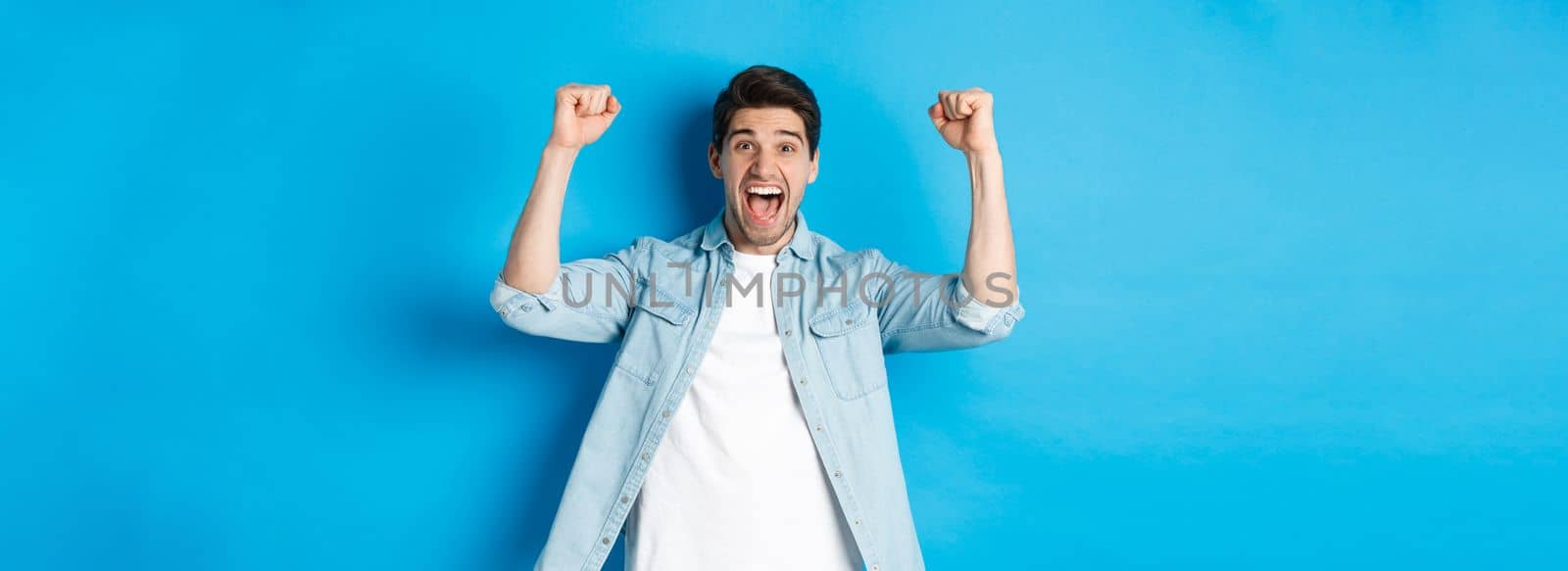 Happy attractive man triumphing, screaming yes and raising hands up to celebrate win, achieve goal, standing against blue background.