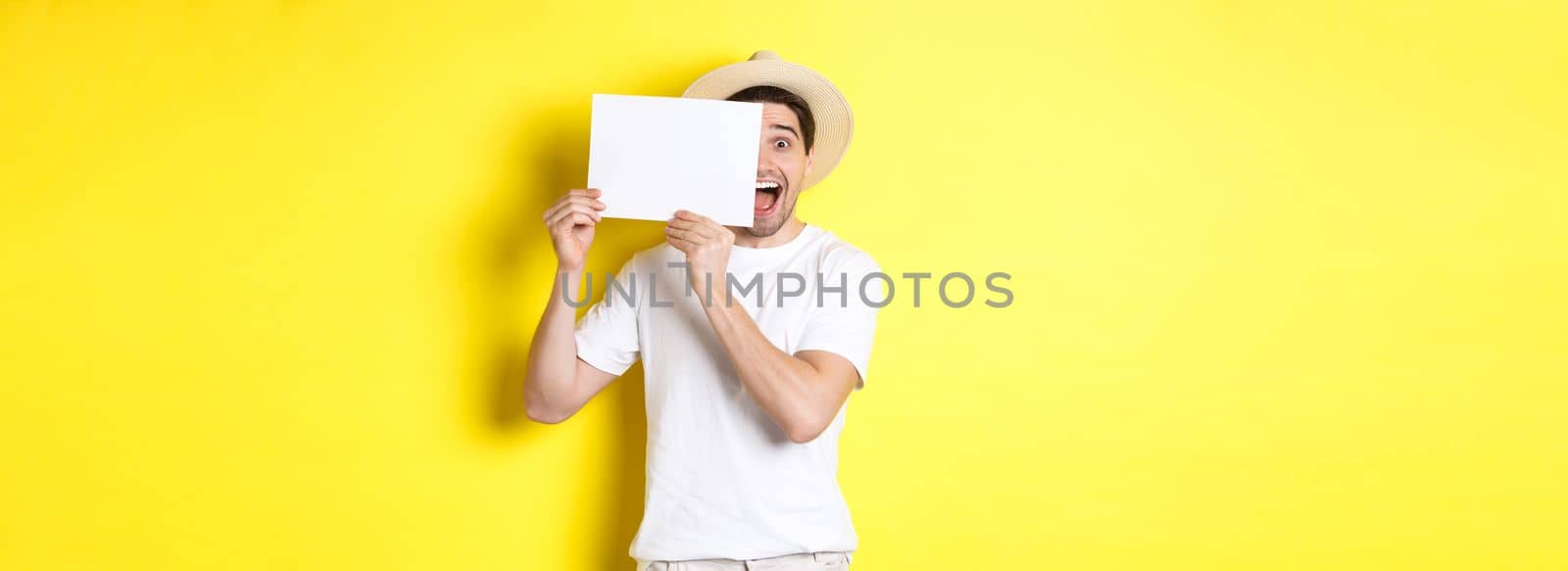 Excited tourist on vacation showing blank piece of paper for your logo, holding sign near face and smiling, standing against yellow background.