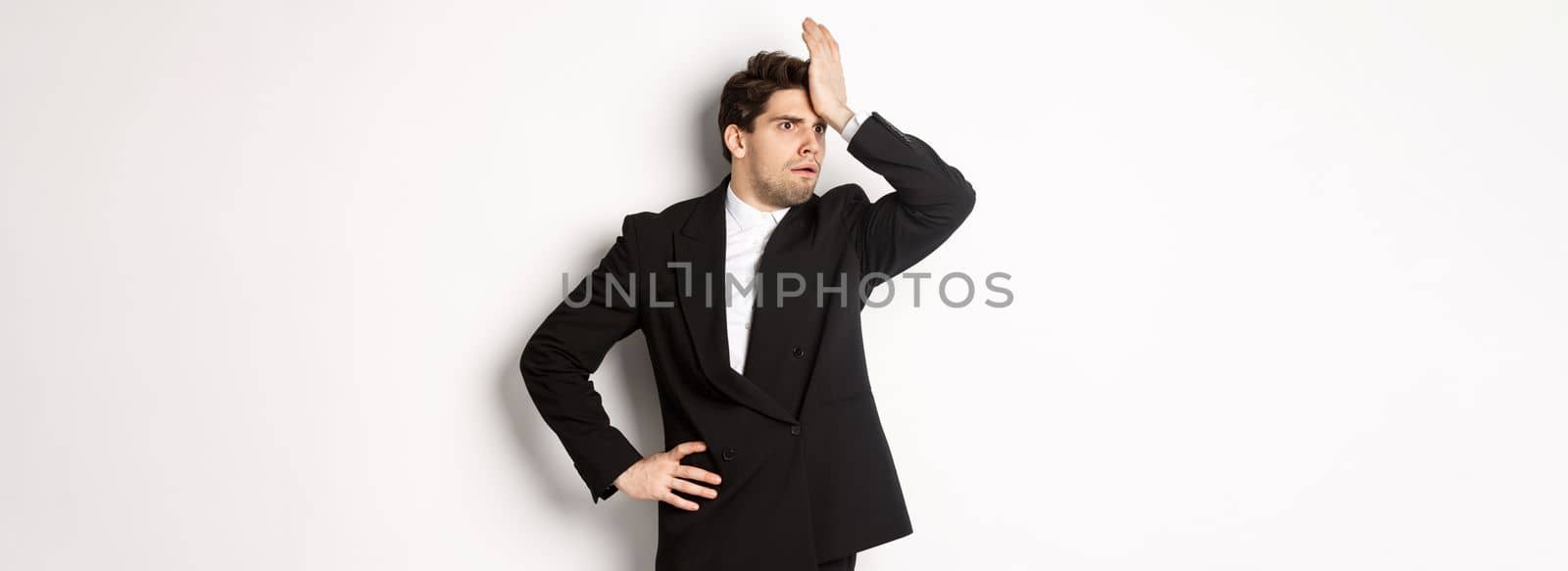 Portrait of shocked and distressed businessman in black suit, slap forehead and looking anxious, standing troubled over white background by Benzoix