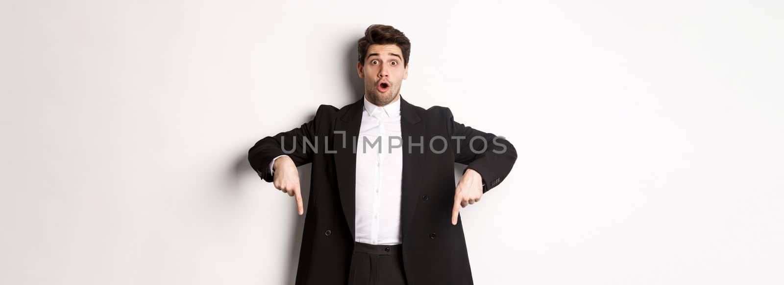 Portrait of surprised and excited man in black suit, pointing fingers down and showing christmas advertisement, standing over white background.