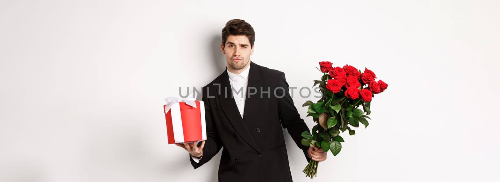 Concept of holidays, relationship and celebration. Handsome and confident man in black suit, going on a date, holding bouquet of roses and present, standing against white background by Benzoix