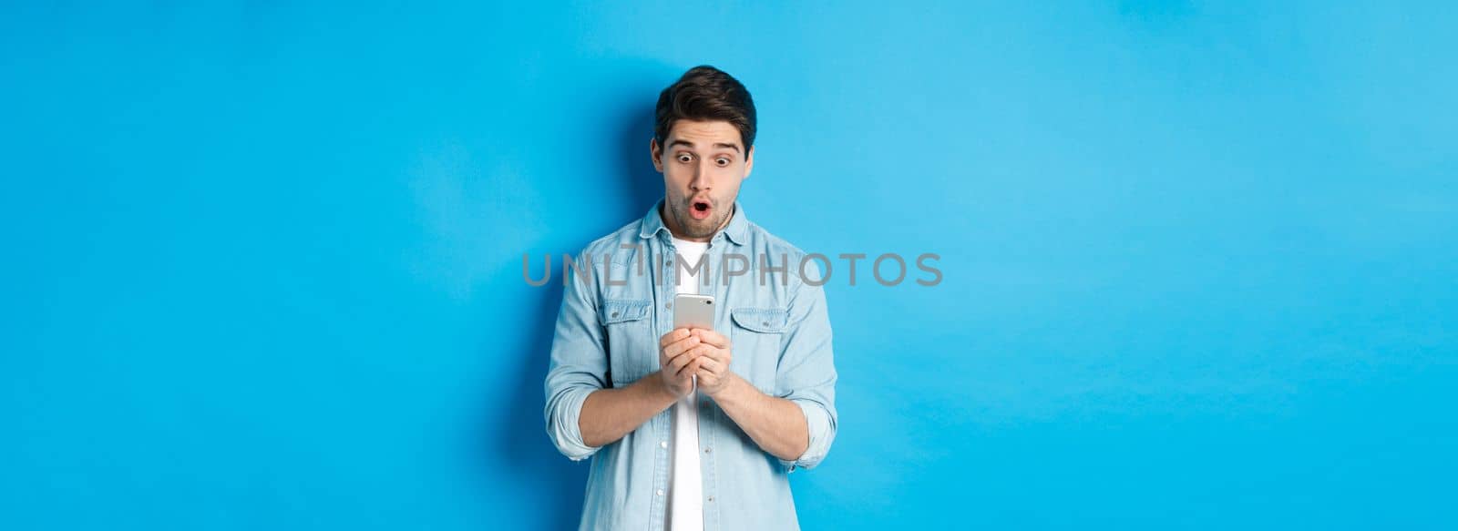 Man looking amazed while checking promo on smartphone, looking surprised at phone, standing against blue background.