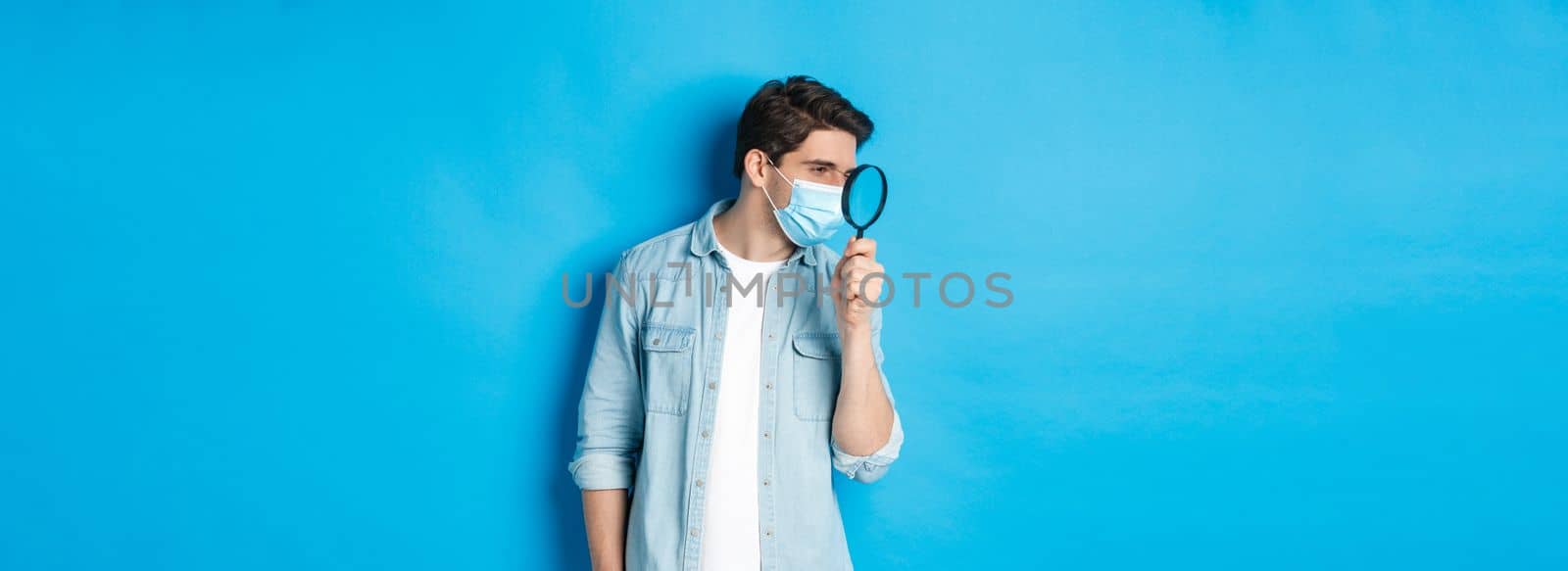 Concept of coronavirus, social distancing and pandemic. Man in medical mask searching for something, looking left through magnifying glass, studying copy space, blue background.