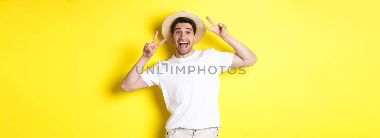 Concept of tourism and vacation. Happy male tourist posing for photo with peace signs, smiling excited, standing against yellow background.