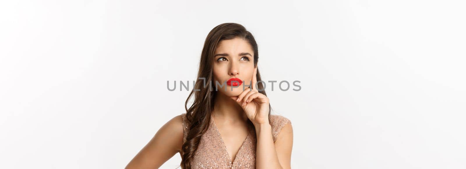 Concept of New Year celebration and winter holidays. Close-up of elegant woman with red lips and dress, looking at upper left corner and thinking, standing over white background.