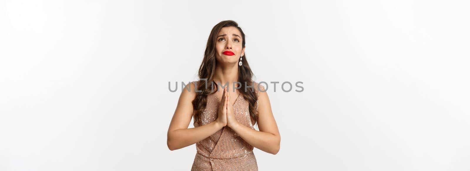 Celebration and party concept. Hopeful young woman begging for help, looking up and praying, pleading God, standing in glamour dress over white background.