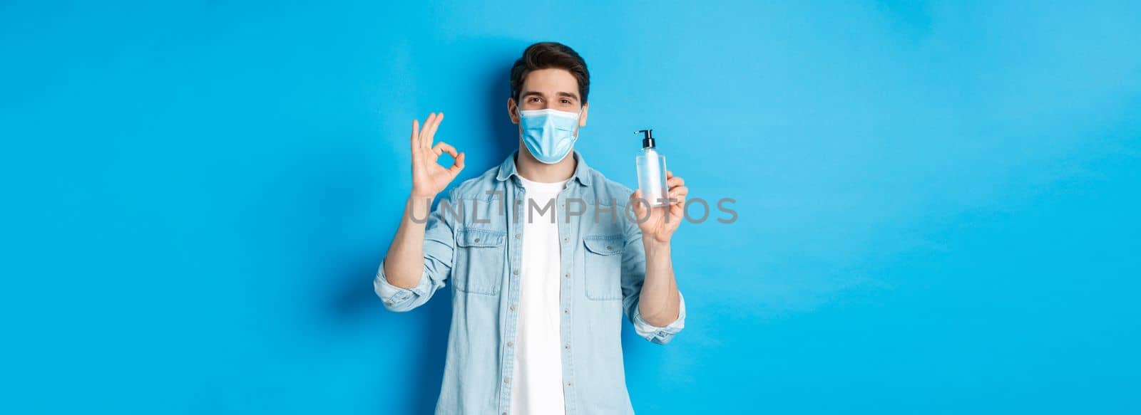 Concept of covid-19, pandemic and social distancing. Satisfied young man in medical mask recommending hand sanitizer, showing ok sign and antiseptic, standing against blue background.