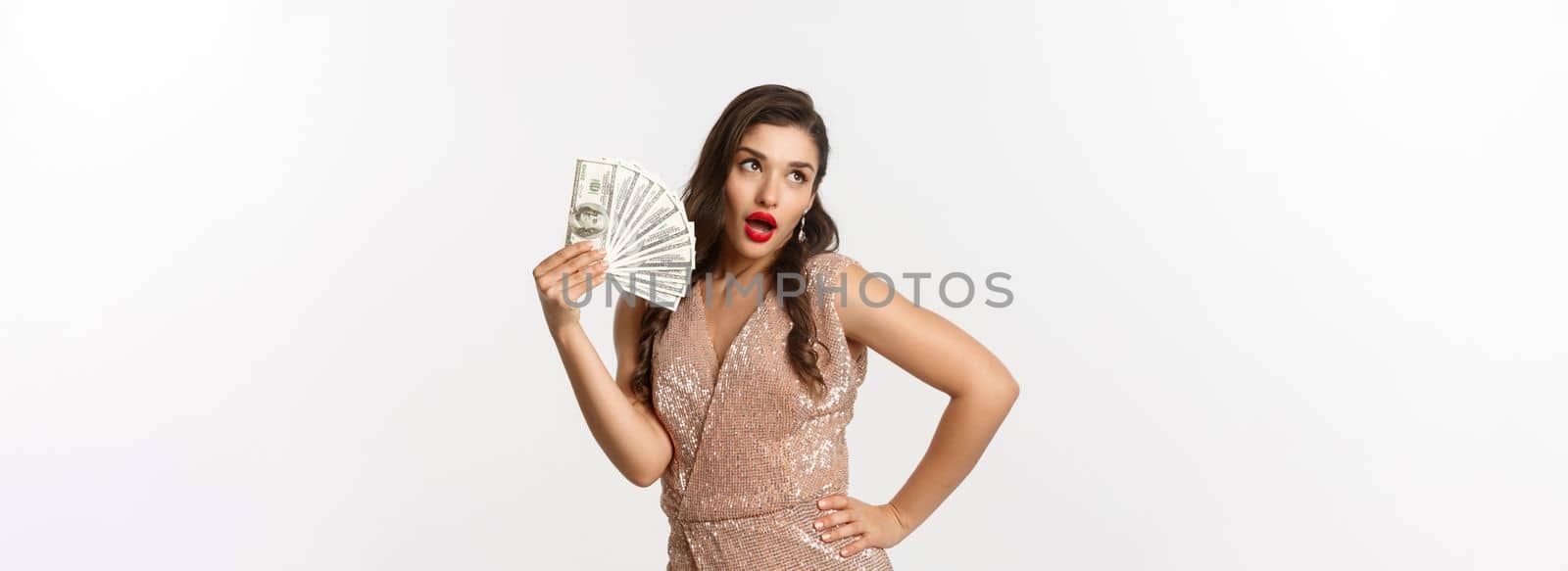 Shopping concept. Successful attractive woman in elegant dress showing dollars, bragging with money, standing over white background by Benzoix
