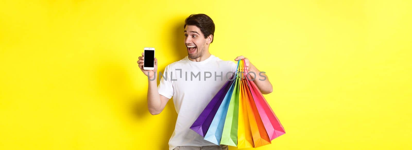 Young man holding shopping bags and showing mobile phone screen, money application, standing over yellow background by Benzoix