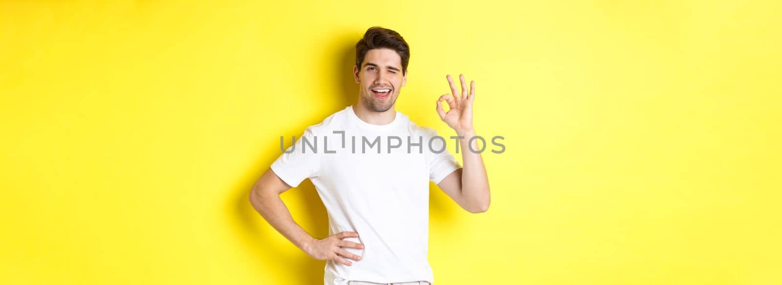 Confident handsome man winking, showing okay sign in approval, like something good, standing over yellow background.