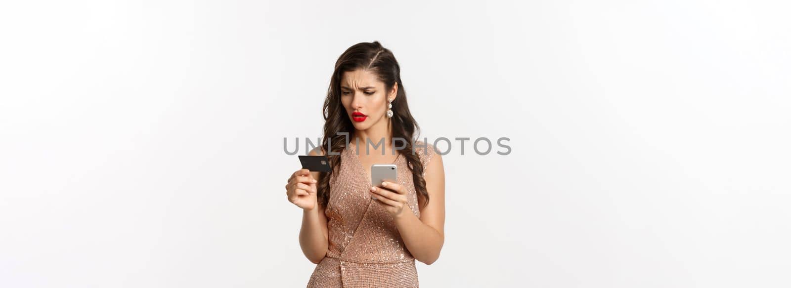 Online shopping and holidays concept. Stylish woman in elegant dress, looking confused at credit card while paying on mobile phone, white background.