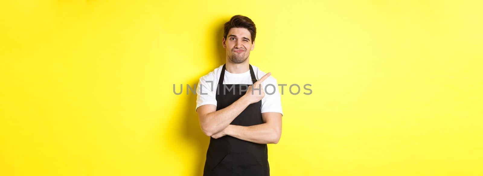 Displeased barista in black apron pointing finger at upper left corner, grimacing skeptical, dislike something, standing against yellow background by Benzoix