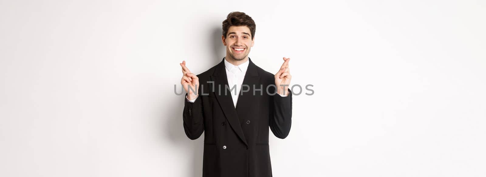 Hopeful and optimistic young businessman in suit, smiling while crossing fingers for good luck and making wish, standing over white background.