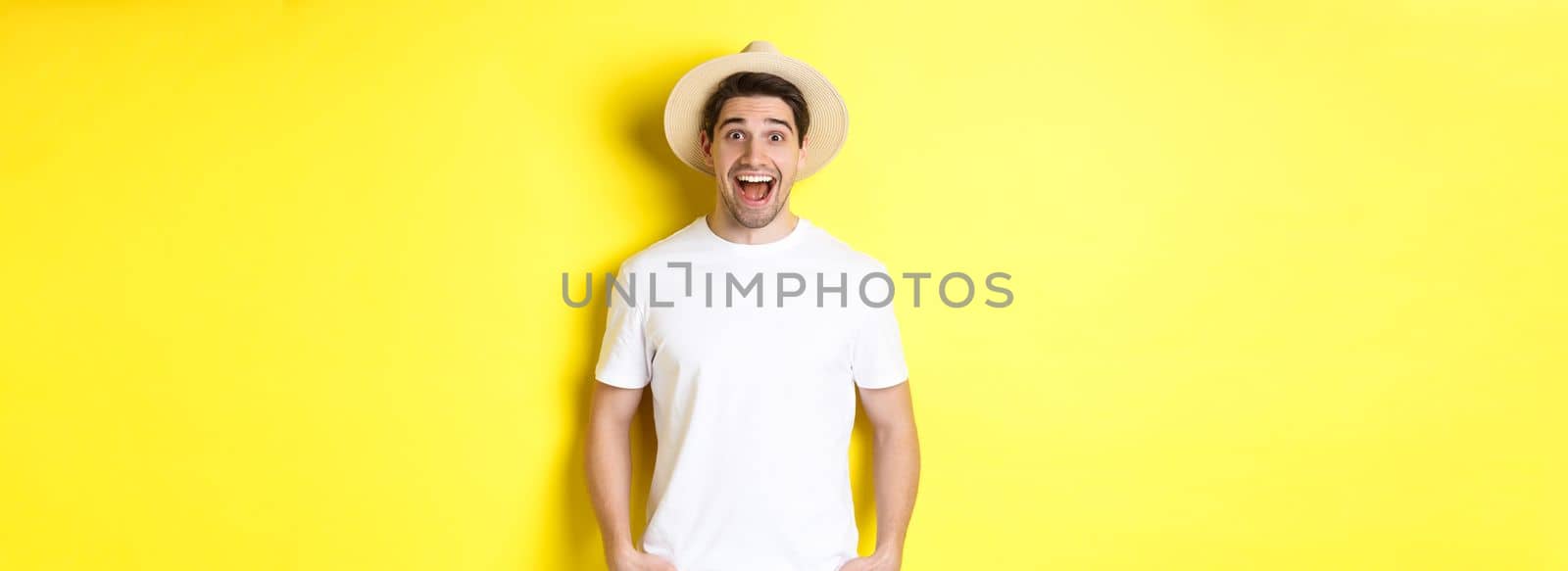 Surprised man tourist in straw hat looking happy, react amazed to travel agency advertisement, standing over yellow background.