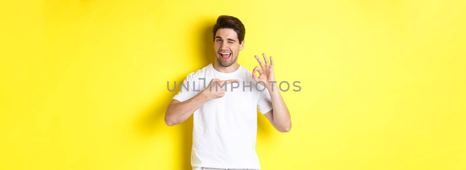 Image of handsome young man approve something, showing okay sign and winking, standing against yellow background by Benzoix
