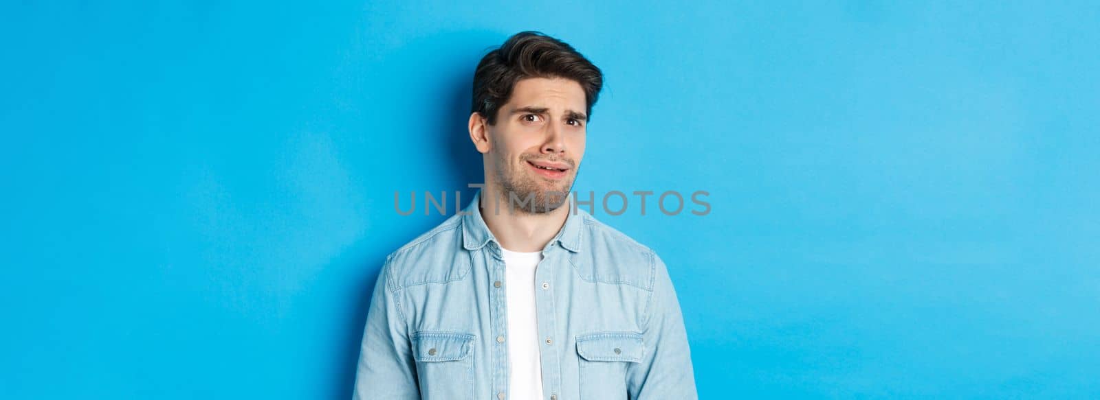 Confused and uncomfortable man looking at something strange or creepy, cringe from bad advertisement, standing over blue background.