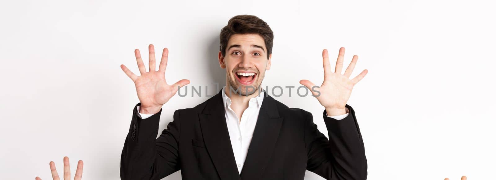 Close-up of handsome businessman in black suit, smiling amazed, showing number ten, standing over white background.