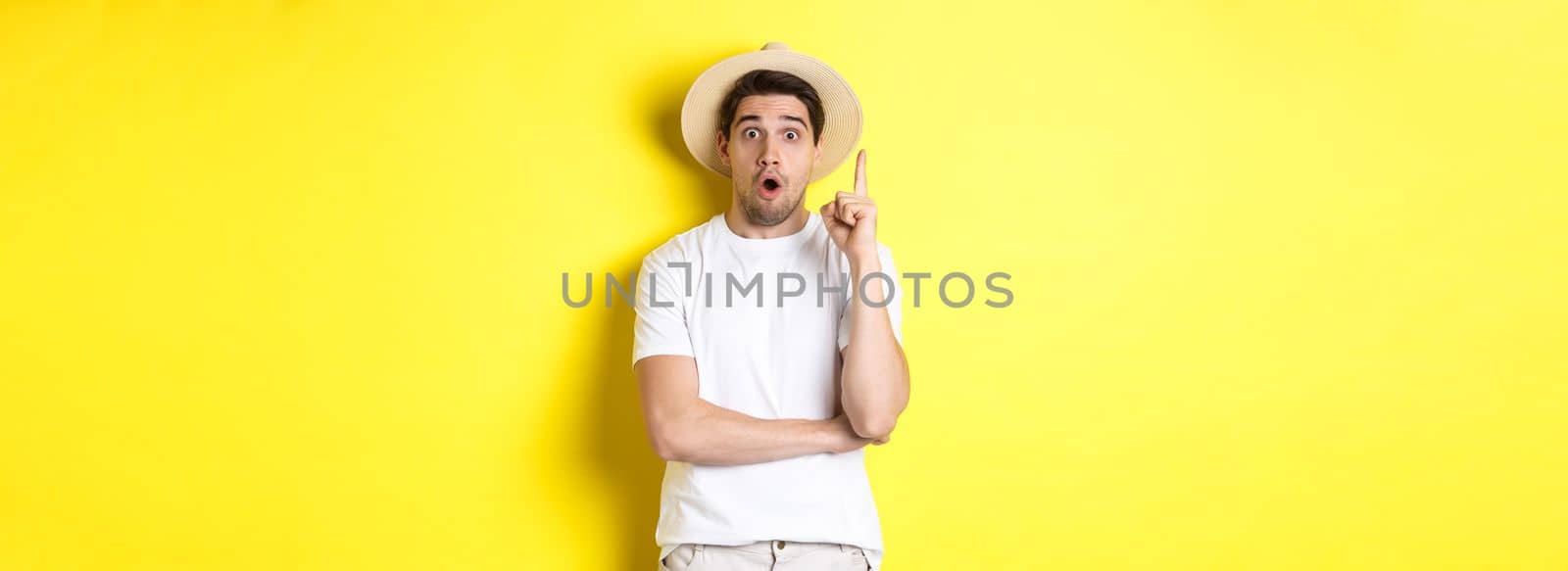 Portrait of young man in straw hat having an idea, raising finger eureka sign, making suggestion, standing over yellow background by Benzoix