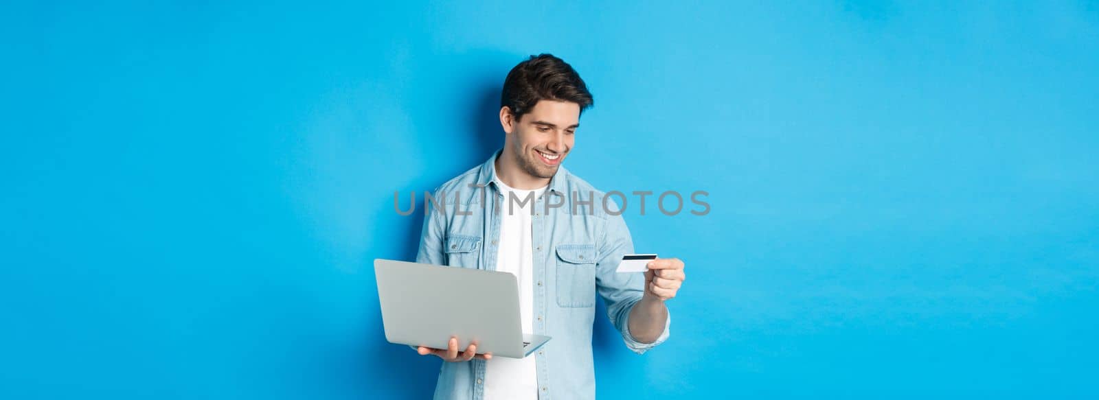 Young man order online, holding credit card and laptop, shopping in internet, standing against blue background.