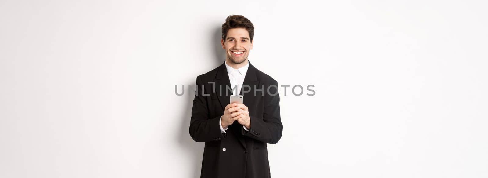 Portrait of successful, handsome businessman in suit, writing message on smartphone and smiling, standing over white background.