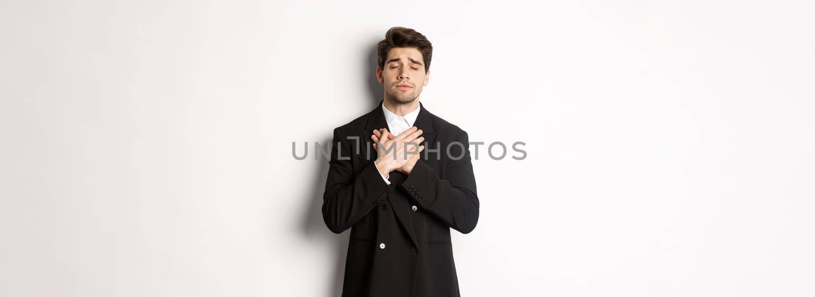 Portrait of dreamy and heartfelt bearded man in suit, close eyes and hold hands on heart, remember something, standing nostalgic over white background.