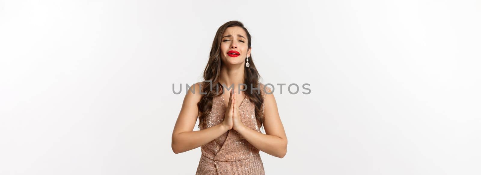 Celebration and party concept. Desperate woman in glamour dress begging for something, need help and holding hands in pray, crying and pleading you, standing over white background.