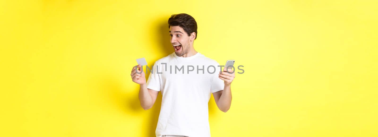 Surprised guy holding smartphone and credit card, online shopping on black friday, standing over yellow background.